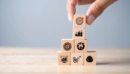 A hand stacks wooden blocks with icons, creating a pyramid-like structure on a wooden surface, against a grey backdrop