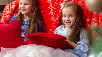 Two sisters in festive dresses sitting in the bedroom decorated with a Christmas tree and a garland