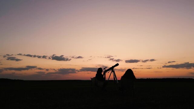 A couple silhouetted against the sunset sky watching space through a telescope