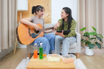 Two young adult woman living together with relationship concept. Asian woman couple relax lifestyle playing guitar on sofa life moments at home. Young lesbian female are playing music.