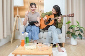 Two young adult woman living together with relationship concept. Asian woman couple relax lifestyle playing guitar on sofa life moments at home. Young lesbian female are playing music.	