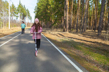 Little girl riding scooter while walking with her grandmother in the forest on a sunny day