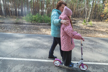 Little girl riding scooter while walking with her grandmother in the forest on a sunny day