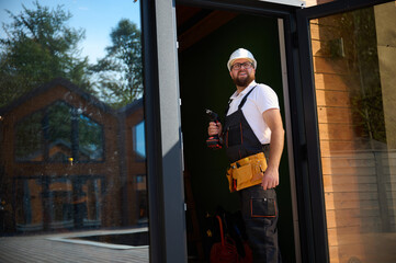 Construction worker installing door in a new house construction site. Man in working attire install front door. Builder in uniform with screwdriver adjusting front glass door in new beautiful house.