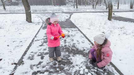 Two little girls in colorful clothes play outdoors during a snowfall.