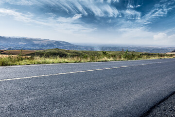 Empty asphalt road and distant rolling hills with mountains under blue sky.