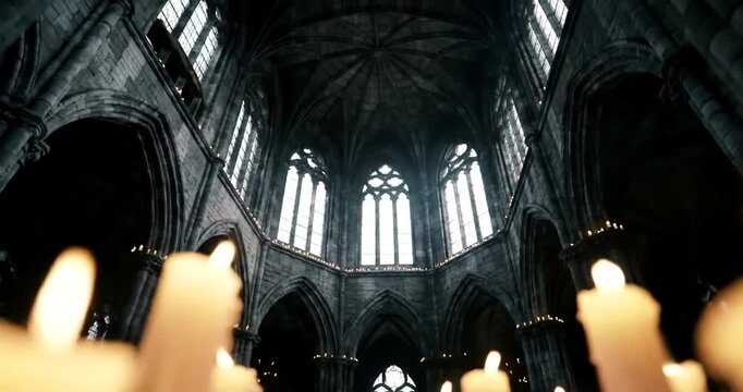 Dimly lit interior of a gothic cathedral with tall arched windows and numerous lit candles in the foreground.