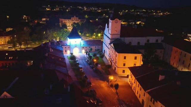 Drone footage of Levoča, Slovakia, at night, featuring a beautifully lit church with twin towers and a glowing green gate, surrounded by historic buildings, UNESCO