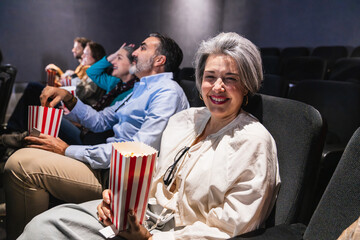 Mature woman smiling and holding popcorn while watching a movie at the cinema with friends, enjoying a leisure activity