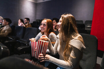 Young women laughing and enjoying popcorn while watching a movie together in a cozy cinema, sharing moments of fun and happiness
