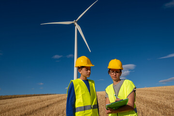 Renewable energy engineer checking clipboard while guiding student near wind turbines,...