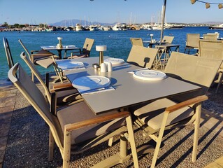 Serene waterfront café with yellow chairs and tables overlooking a calm sea, framed by boats and distant buildings under a clear sky. Metaphorically, it symbolizes a haven of rest, connection.