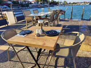 Serene waterfront café with yellow chairs and tables overlooking a calm sea, framed by boats and distant buildings under a clear sky. Metaphorically, it symbolizes a haven of rest, connection.