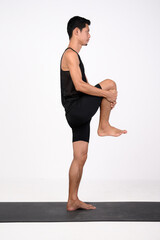 A male runner in black athletic wear performs a standing knee-to-chest stretch on a mat. A man...