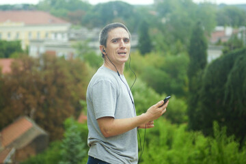 Fototapeta premium Middle aged man wearing earphones with mobile device, cheerfully singing while walking outside. Сoncept of karaoke mood, joyful lifestyle, digital entertainment, creativity, mental health, outdoor fun
