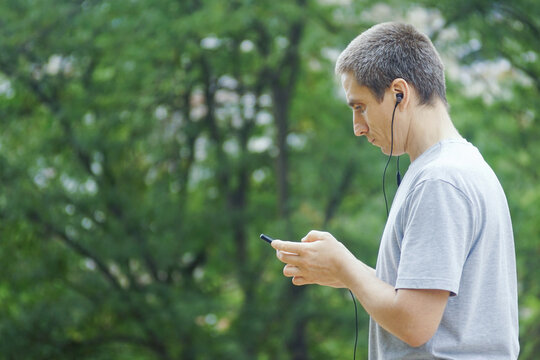 Millennial man listening to educational podcast or audiobook content on mobile phone with earphones in green park. Concepts of lifelong learning and personal development through audio media