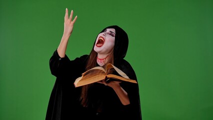 A vampire witch in a black cloak casts spells over a magic book against the green background of a...