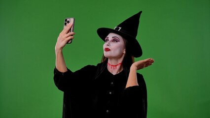A witch wearing vampire makeup, dressed in a black cloak and hat, talks on a video call on her...
