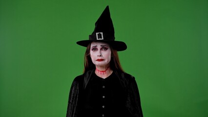 A witch with vampire makeup, dressed in a black cloak and hat, looks sadly into the camera