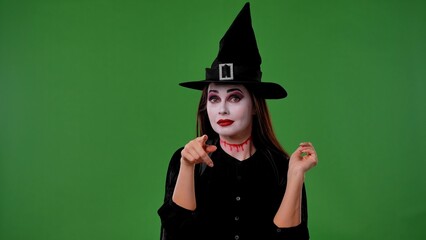 A witch with vampire makeup, in black cloak and hat, points her index finger at the camera