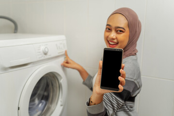 A woman is joyfully showing off her smartphone while standing next to a washing machine