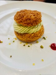 Elegant choux pastry filled with pistachio cream, artfully presented on a white plate with raspberry coulis dots and pistachio crumbles.