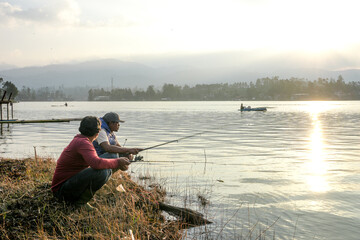 Two Men Crouch On The Lakeshore, Casting Their Fishing Rods Into The Calm Water As The Sun Sets