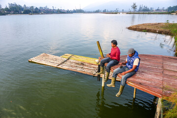 Two Men Sit On A Wooden Dock, Fishing With Rods In A Calm Lake With A Distant Village