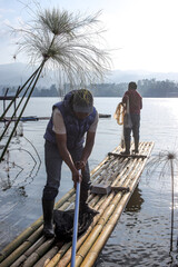 Two Men On A Bamboo Raft Prepare To Fish On A Peaceful Lake With Mountains In The Background