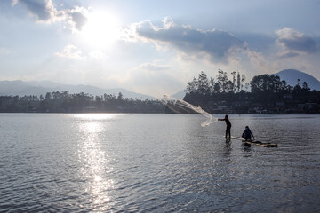 Two Fishermen On Bamboo Raft Casting Net Into The Serene Lake