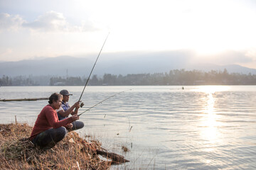 Two Men Crouching On the Grassy Shore Of Lake Fishing During Sunrise