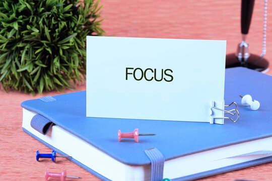 A card displaying the word focus is attached to a blue notebook on a desk surrounded by colorful push pins and a small green plant, suggesting a workspace atmosphere