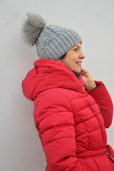 Portrait of pretty Caucasian woman in red winter jacket and grey knitted hat with fur pompom on white background. Women's warm street clothes.