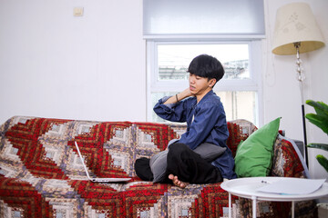 Young Asian Man With Strained Neck Sitting On Living Room Sofa Taking break From Working On Computer