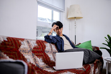 Anxious Young Male Working From Home Looking Upset While Sitting On Couch With Hand On Head
