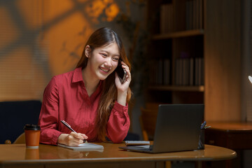Young Asian businesswoman talking on mobile phone and taking notes in notebook while working late...