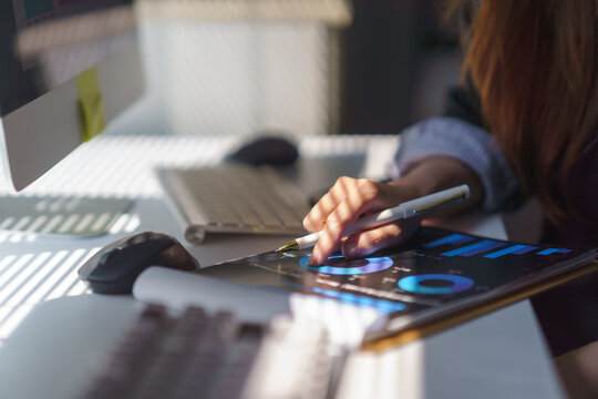 Businesswoman pointing at financial charts on a digital tablet, analyzing data in a modern office filled with natural sunlight - Powered by Adobe