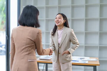 Two Asian businesswomen smiling and shaking hands after successfully closing a deal in a modern office filled with natural light