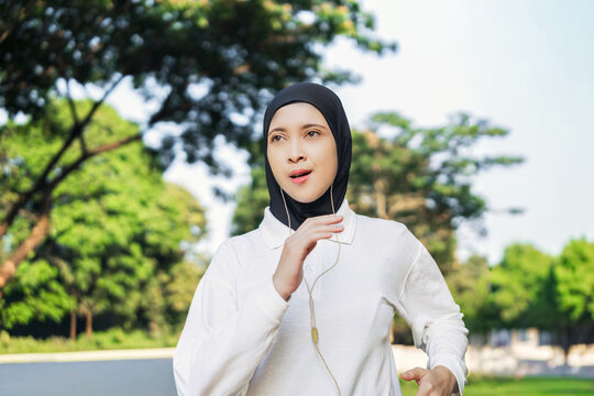 Asian woman in a hijab jogging outdoors, symbolizing fitness, determination, and active living. The background setting includes green park under bright sunlight, conveying health and mindfulness. - Powered by Adobe