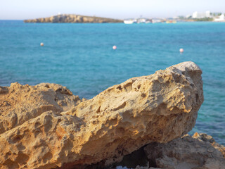 View from behind a big yellow porous rock over a beautiful blue Mediterranean sea, Nissi beach, small island and hotel buildings in the background in Ayia Napa, Cyprus.