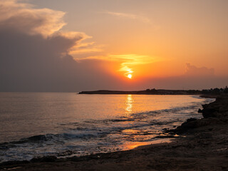 Orange orb of a sun setting above the Mediterranean sea in Ayia Napa, view from the rocky shore over the curvy shore, waves and sunset glade on the water surface. Picturesque evening landscape concept