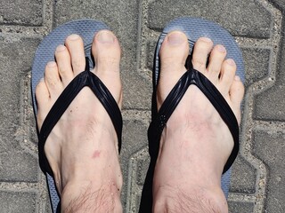 Bare feet in worn black flip-flops on a sunlit pavement, symbolizing a carefree summer stroll....