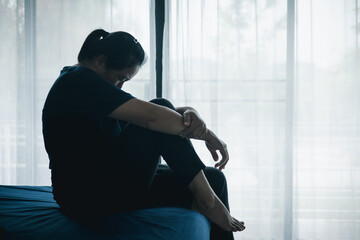 A stressed woman sitting alone on bed in dark room, holding her head down, symbolizing depression,...