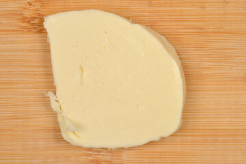 A single, thick slice of fresh white mozzarella cheese rests on a light brown wooden cutting board, photographed from a top-down view