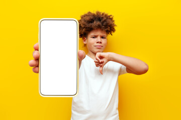 dissatisfied african american boy showing blank smartphone screen and dislike on yellow isolated...