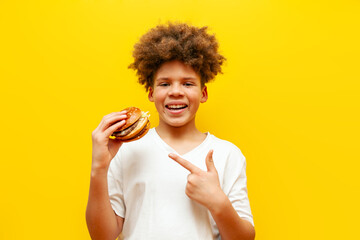 cheerful curly african american boy showing burger on yellow isolated background, hungry teenager...