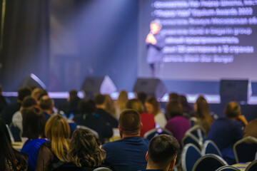 Audience listening to presenter during business conference, with focus on attendees and stage.