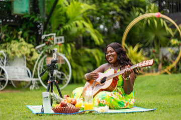 Music and social media, African woman sitting on a picnic mat in the park, playing acoustic guitar...