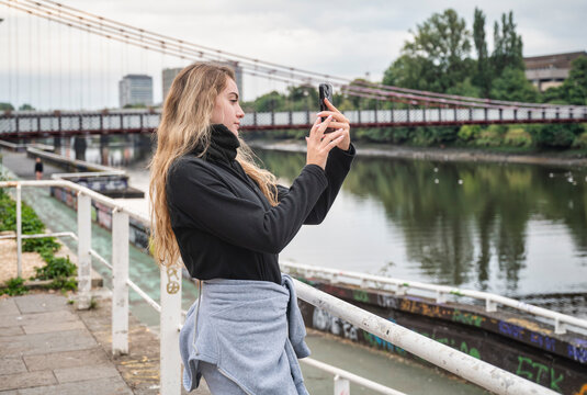 Young woman taking photo with smartphone by river and bridge in city during a cold vacation day - Powered by Adobe