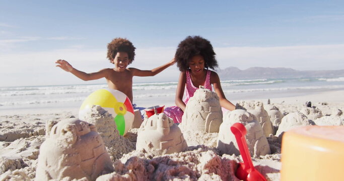 Kneeling siblings molding sandcastles at sandy beach edge, with red shovel and orange bucket